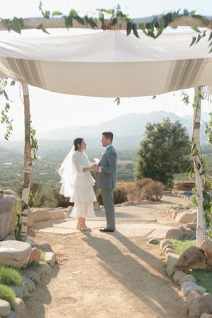 Couple exchanging vows under a white canopy chuppah with mountain views at an outdoor wedding ceremony.