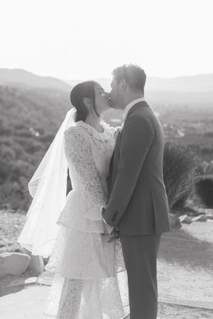 Black and white photo of a bride and groom sharing a kiss outdoors with scenic hills in the background.