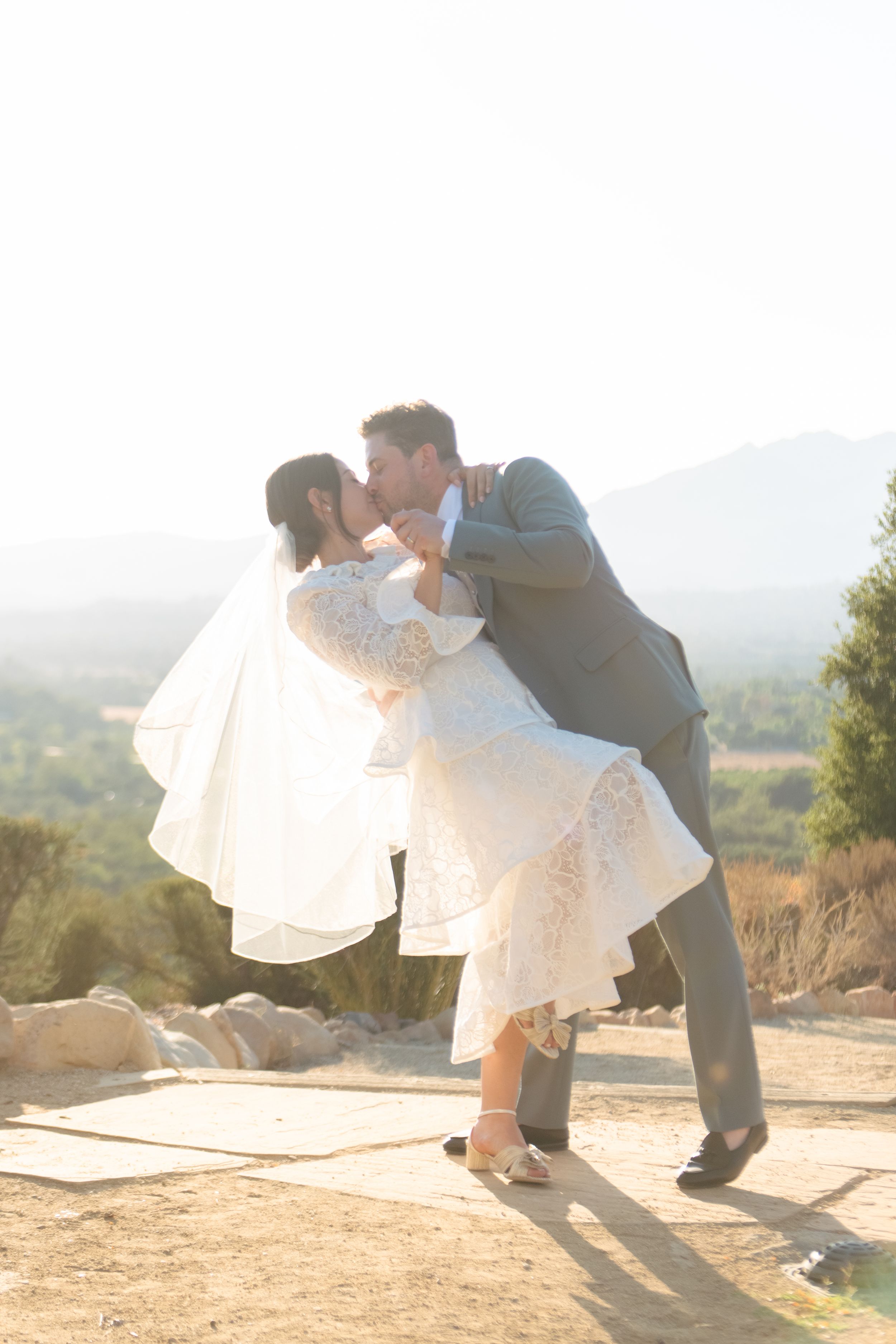 A groom dips his bride for a romantic kiss outdoors, with scenic mountains and golden sunlight in the background.