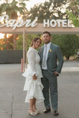 Bride and groom posing in front of Capri Hotel sign at golden hour, she in lace dress, he in gray suit.