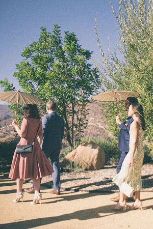 Three elegantly dressed guests walk outdoors holding parasols at a desert garden wedding celebration.