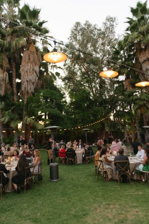 Outdoor evening wedding reception on lush lawn with string lights, pendant lamps, and guests dining under tall palm trees.