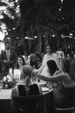 Black and white photo of a bride in lace dress greeting guests at a wedding reception dinner table outdoors.