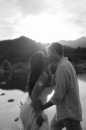 Black and white photo of a couple sharing a kiss at sunset with mountains and a lake in the background.