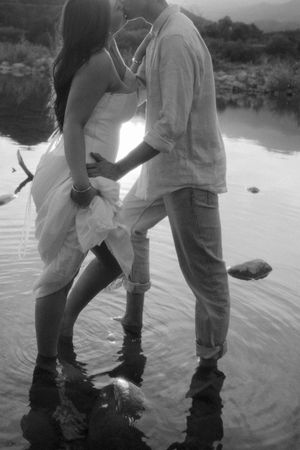 Romantic couple kissing while standing in shallow water, black and white photo, woman in white dress, man in casual attire.
