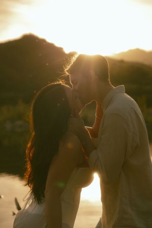 Couple sharing a romantic kiss at golden sunset with mountain backdrop in warm glowing light.