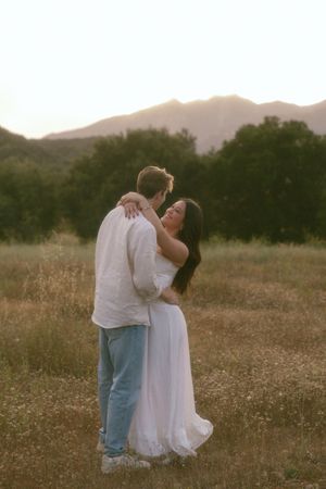 Couple embracing in golden meadow at sunset with mountains and trees in background.