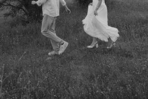 Black and white photo of a couple in white outfits running through a grassy field, capturing a joyful romantic moment.