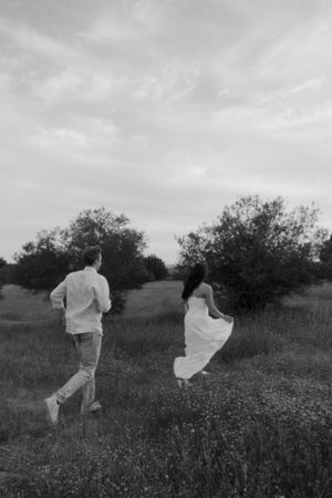 Couple runs through wildflower meadow in black and white photo, woman in white dress, cloudy sky.