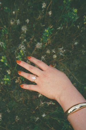 Woman's hand with orange nails, diamond ring, and gold bracelet resting over wildflowers in a green field.