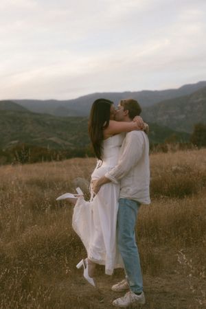 Couple embracing and kissing in golden field with mountain backdrop at dusk.