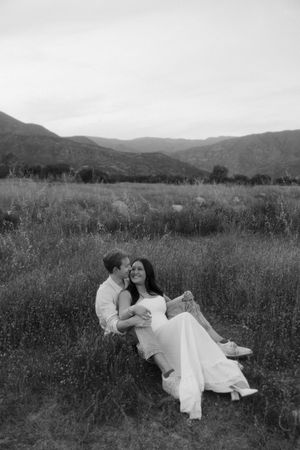 Couple sitting together in meadow laughing with mountain landscape in black and white.