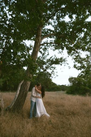 Romantic couple kissing under large oak tree in dry golden field at dusk.