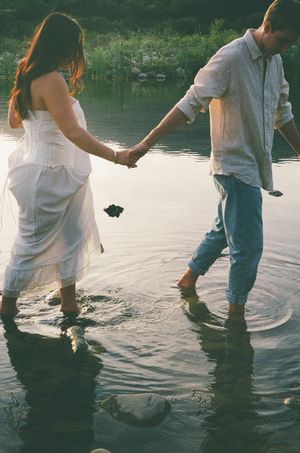 Couple holding hands wading through calm lake water, woman in white dress at sunset.