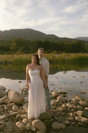 Couple standing on rocky shore by reflective lake with mountain backdrop at golden hour.