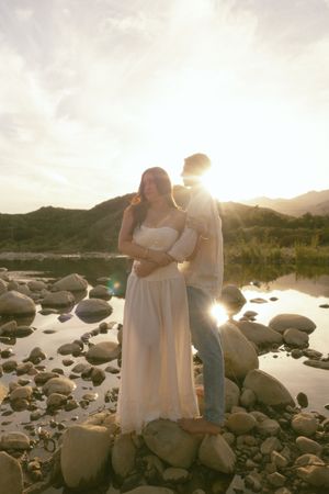 Couple embracing on rocky riverbank at golden sunset with mountains in background, woman in white dress.
