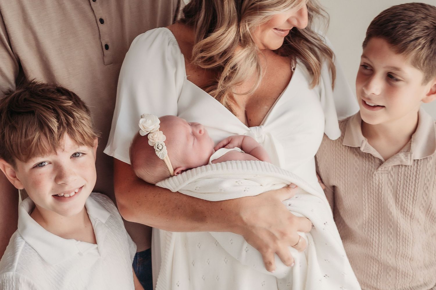 A mother in a white dress holds her newborn baby surrounded by two smiling boys in a bright family portrait.
