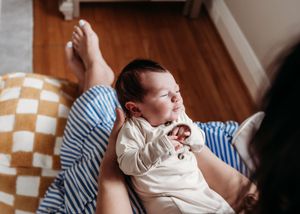 Newborn baby resting peacefully on parent's lap in cozy home setting, wearing white onesie.