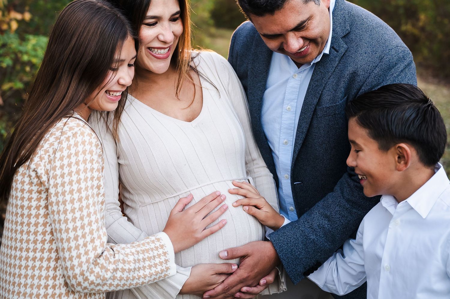 Family gathered around pregnant woman touching her baby bump outdoors in a warm, joyful maternity photo session.