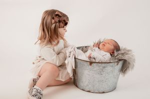 Young girl with bows gazing at sleeping newborn in rustic metal tub.
