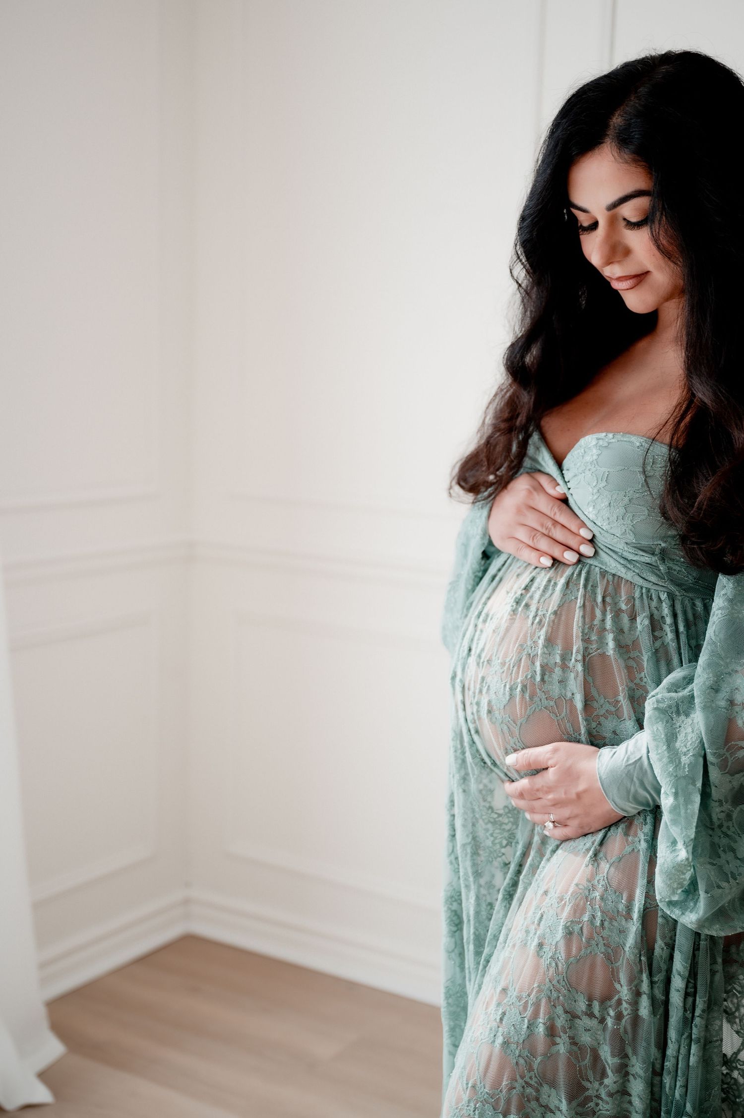 Pregnant woman in green lace gown gently holding her baby bump indoors.