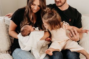 Family of four with parents in black shirts cuddling newborn and toddler girl.