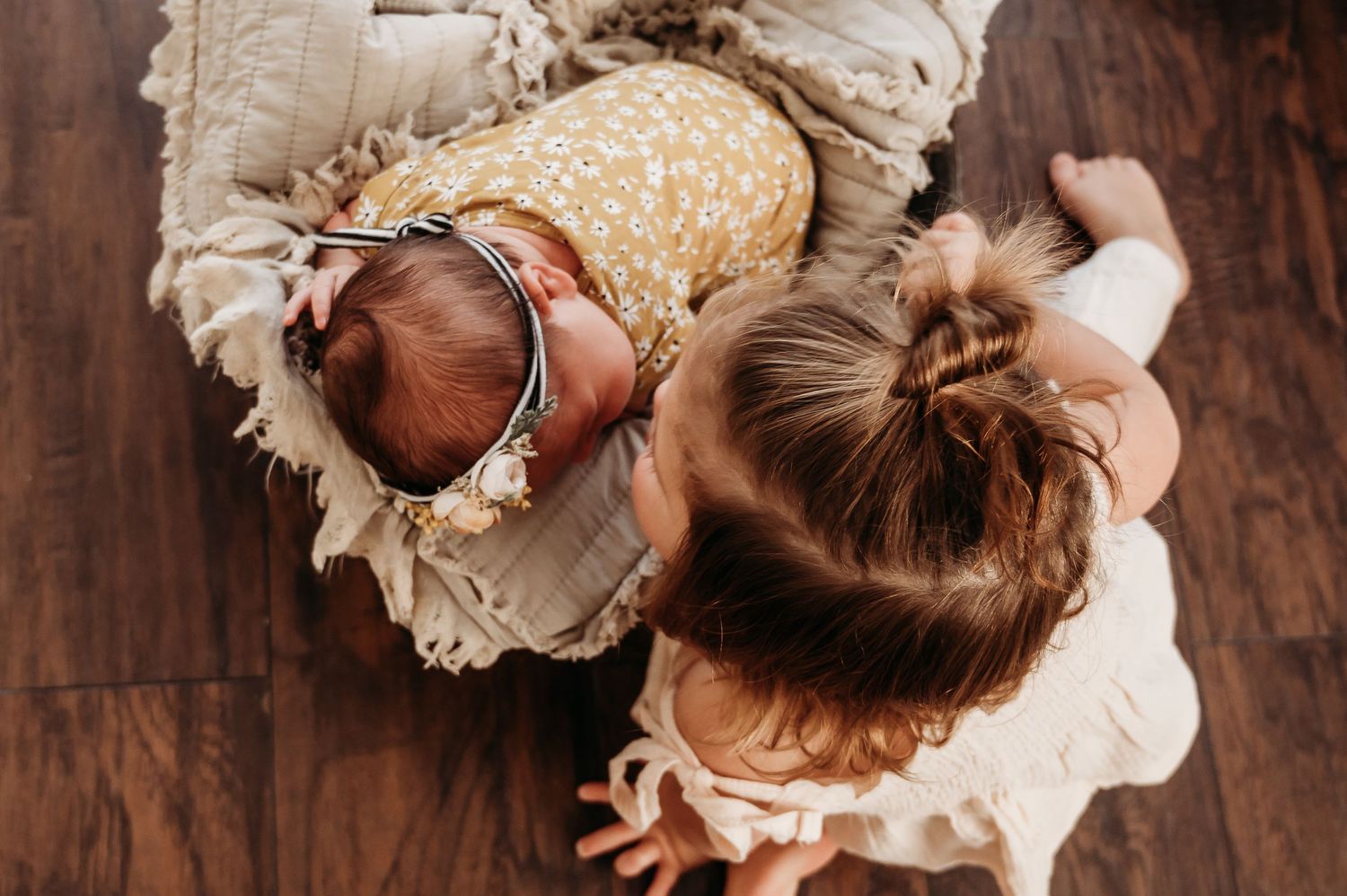 Overhead view of toddler girl leaning over newborn baby on wooden floor.