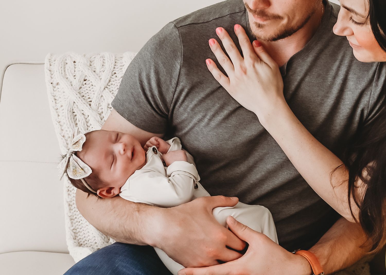 Parents tenderly holding sleeping newborn baby together on white sofa.