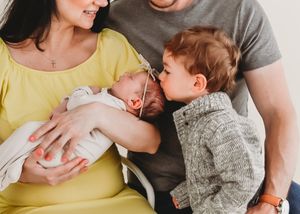 Family with newborn baby, toddler kissing infant sibling held by mother in yellow dress.