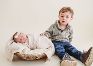 Toddler boy in grey sweater sitting beside sleeping newborn in wicker basket.