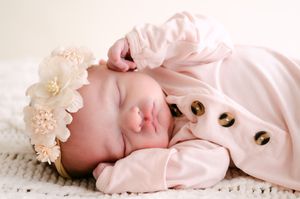 Sleeping newborn baby girl in pink outfit with floral headband on white knit blanket.