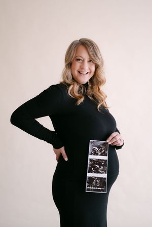 Smiling pregnant woman in black dress holding ultrasound photos against neutral background.