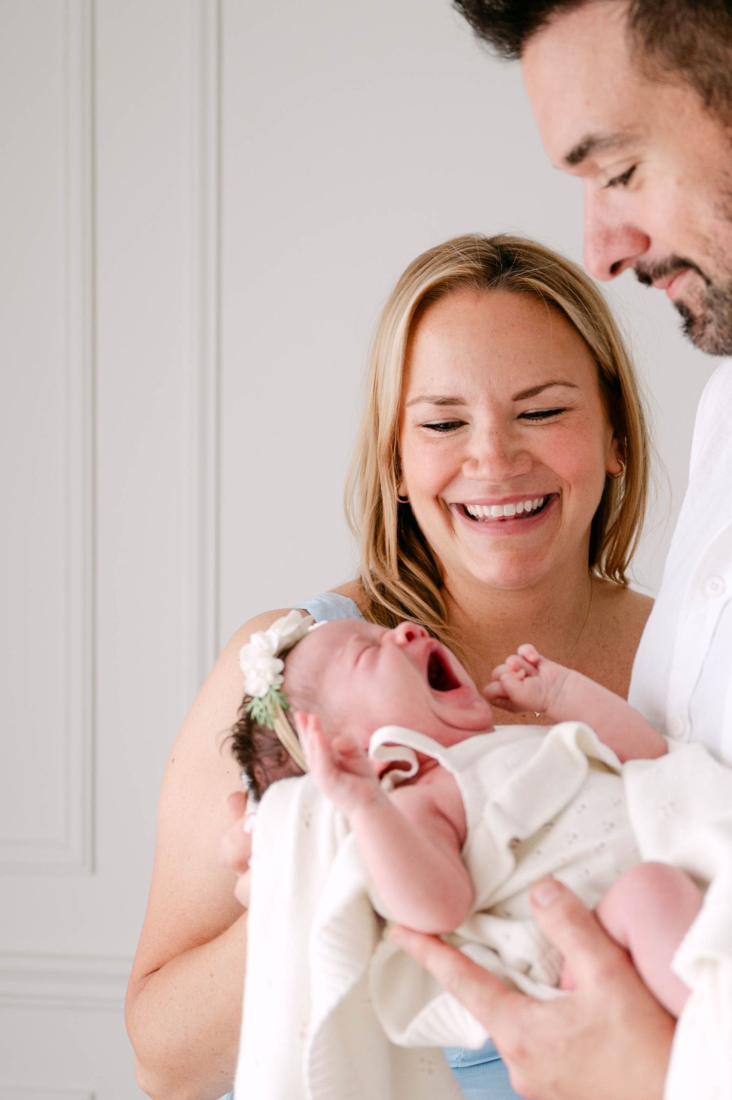 Joyful parents laughing while holding their yawning newborn baby with floral headband indoors.