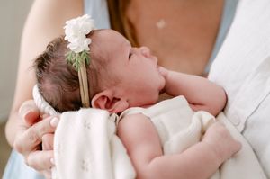 Newborn baby with white flower headband sleeping peacefully in mothers arms wrapped in blanket.