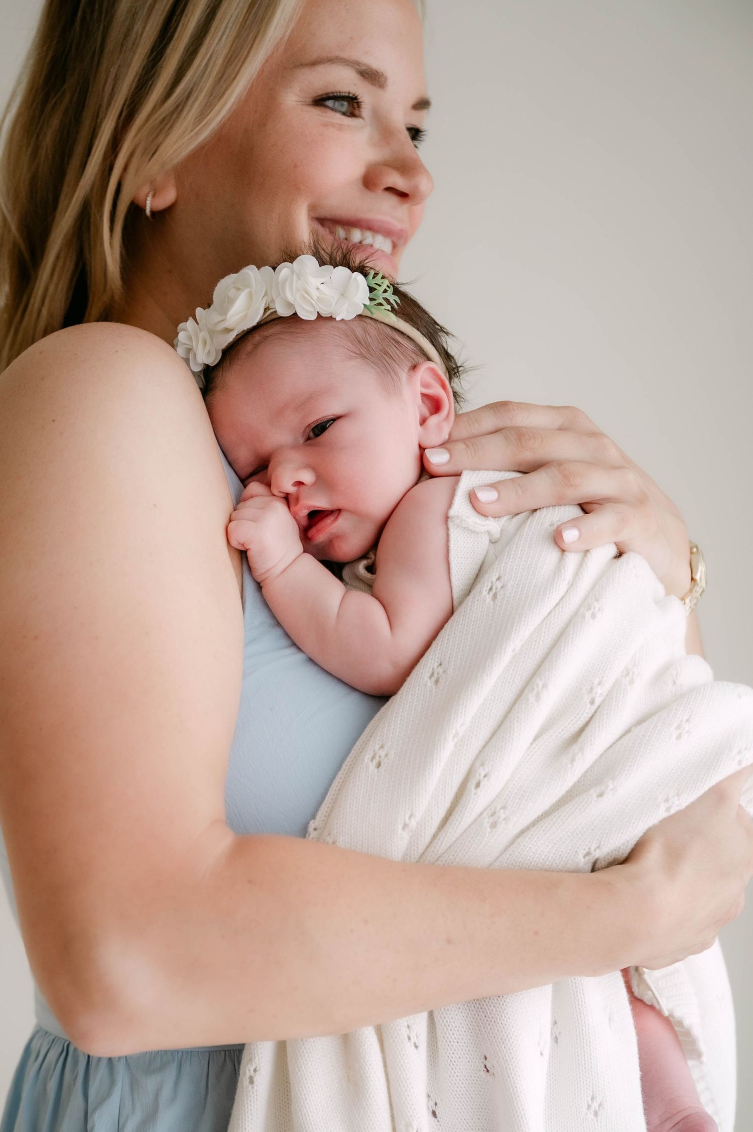 Smiling blonde mother cuddling newborn baby with floral headband wrapped in white swaddle blanket.