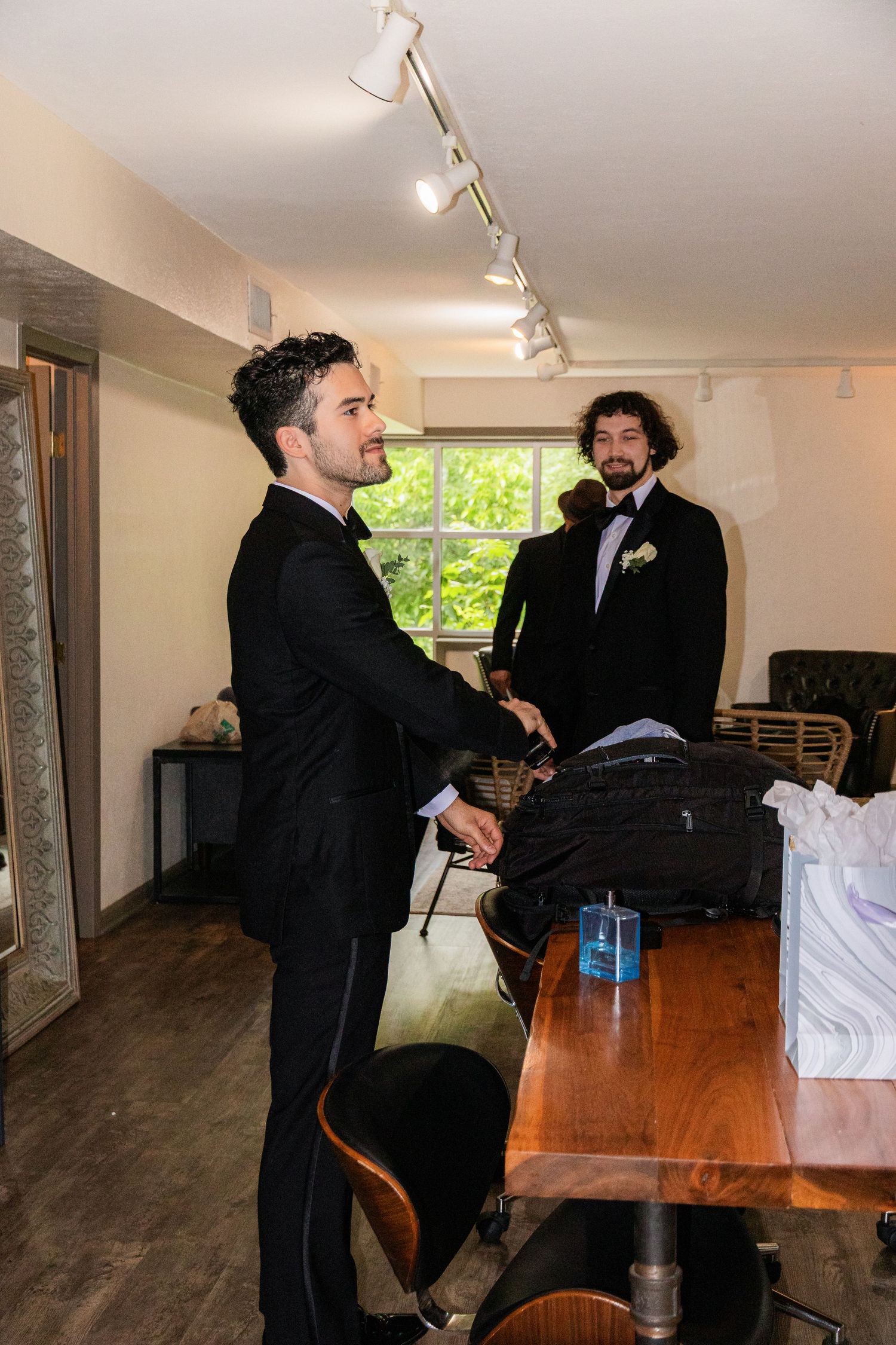 Two men in formal black tuxedos stand in a room, one wearing a boutonniere, preparing for a wedding.