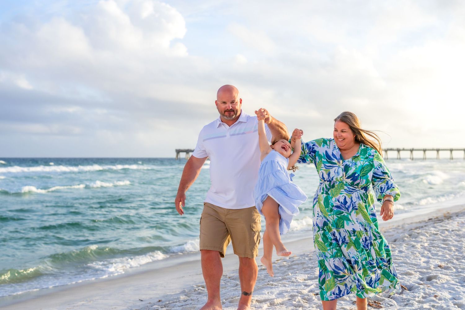 Family walking on a sunny beach, smiling couple swinging child between them with ocean waves in the background.