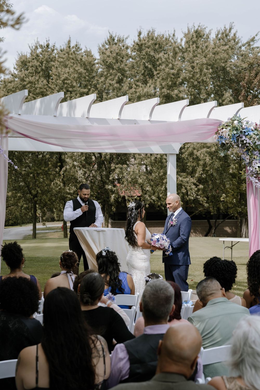 Outdoor wedding ceremony under white pergola with pink draping, bride and groom at altar with guests seated on white chairs.