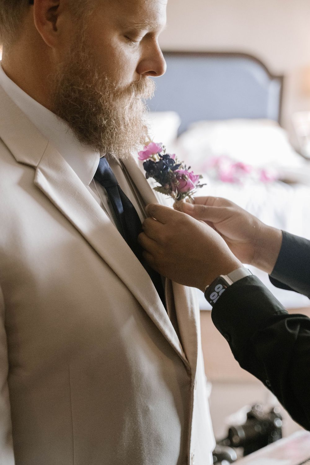 Groom in beige suit having purple floral boutonniere pinned to lapel by groomsman.