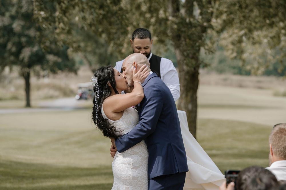 Bride and groom share their first kiss at an outdoor wedding ceremony on a golf course, officiant standing behind them.