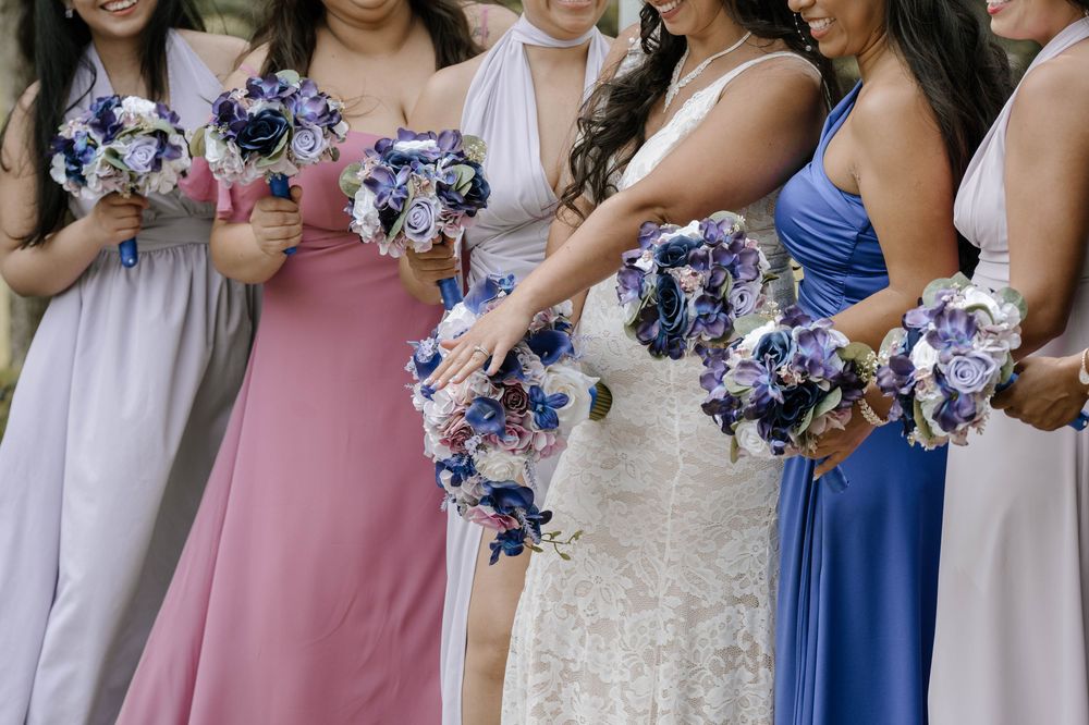 Bride and bridesmaids in pink and blue dresses holding purple and blue floral bouquets at a wedding ceremony.
