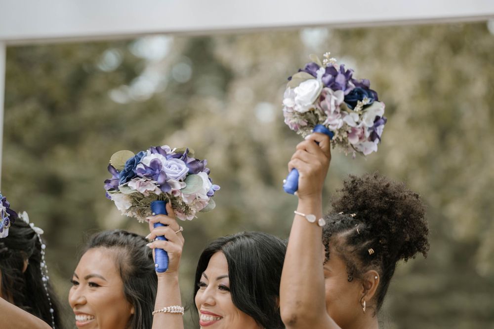 Bridesmaids in blue dresses raising floral bouquets with purple and white flowers at a wedding ceremony.