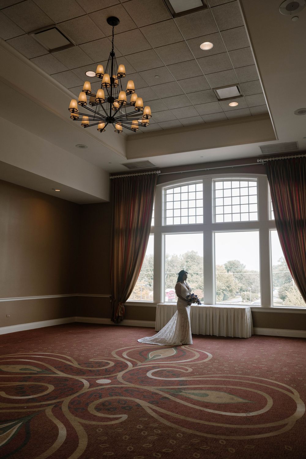 Bride in white gown stands by large window holding bouquet in elegant ballroom with chandelier and ornate carpet.