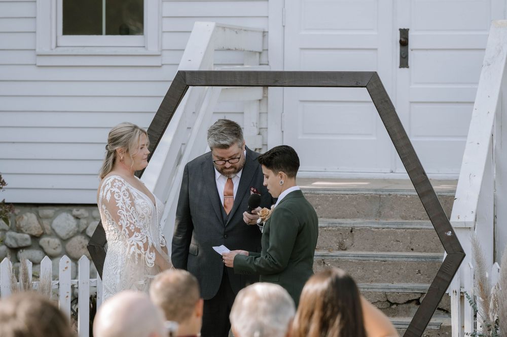 Outdoor wedding ceremony with bride in lace gown, groom, and officiant standing before hexagonal arch backdrop.