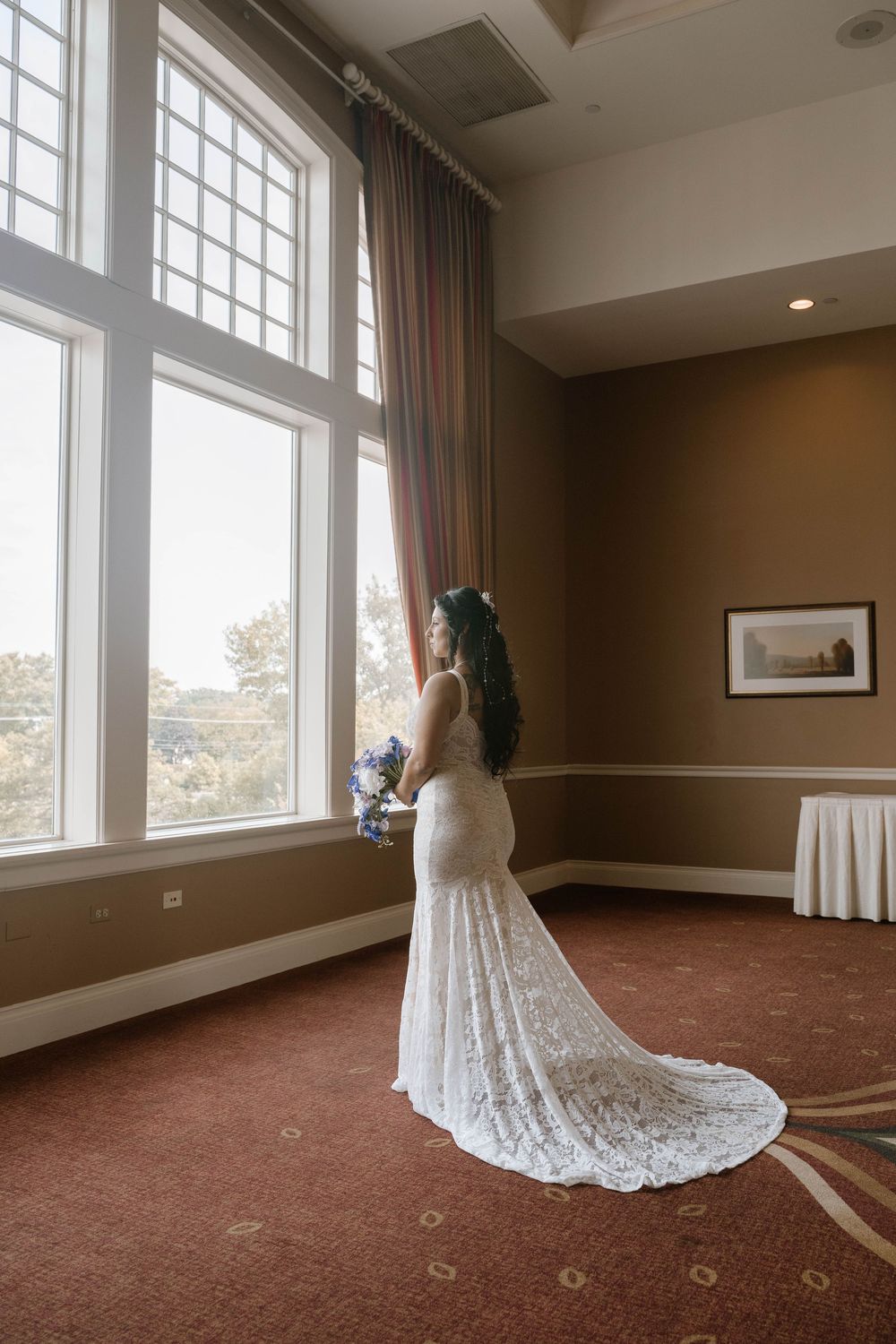 Bride in lace mermaid gown holding bouquet stands by tall windows in elegant room with warm brown walls.