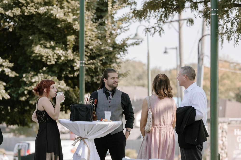 Guests mingle outdoors at a wedding cocktail hour around a high-top table with string lights and greenery.