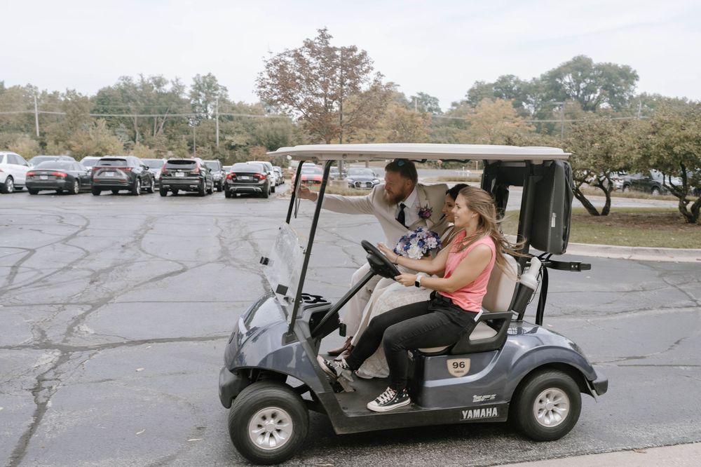 Two people riding a Yamaha golf cart in a parking lot surrounded by trees and parked cars.