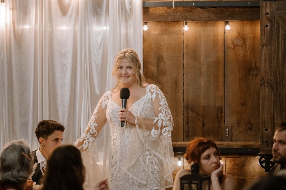 Blonde woman in lace wedding dress giving a speech with microphone at rustic barn wedding reception.
