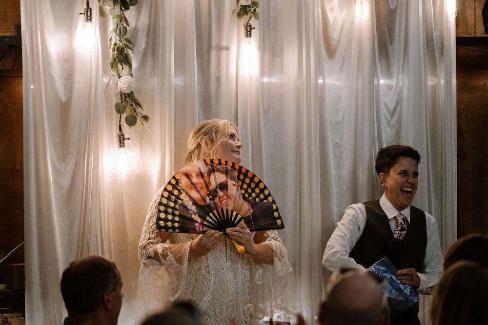 Bride in lace gown holding decorative fan laughing with guest at elegant barn wedding reception.