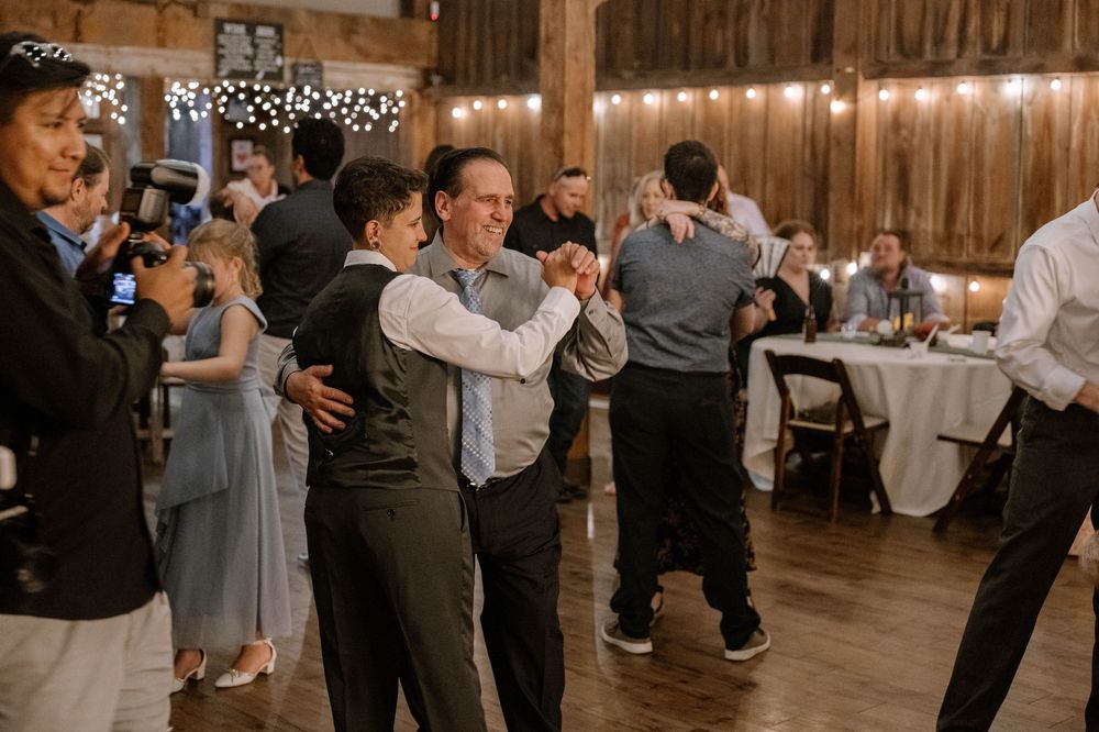 Guests dancing together in a warmly lit rustic barn wedding reception with string lights overhead.